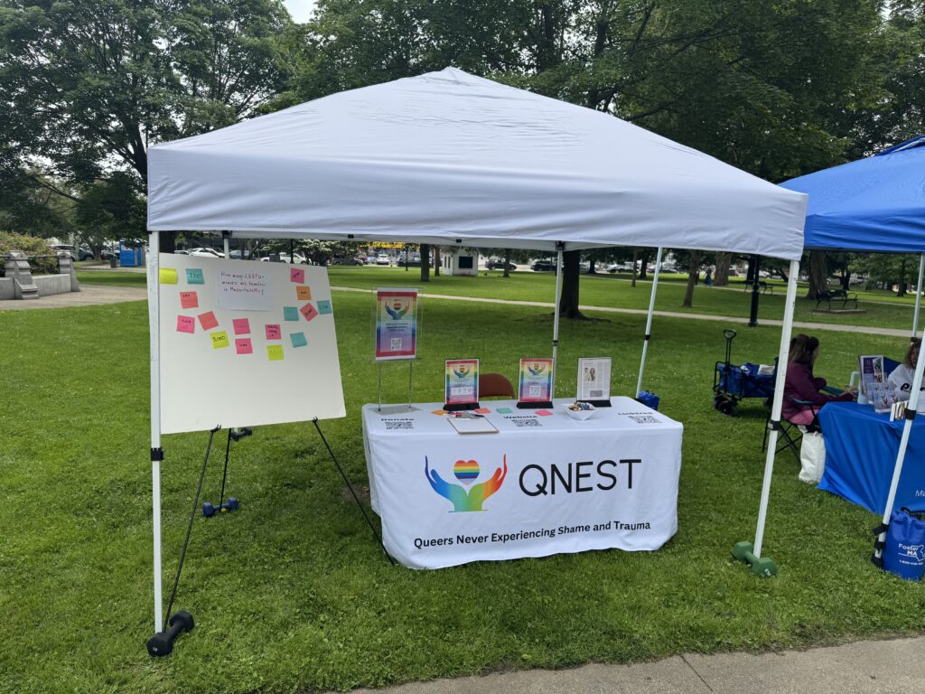 A table under a tent with a white tablecloth displaying QNEST's name and logo along with other displays on the table. Next to the table is a board with post it notes on it.